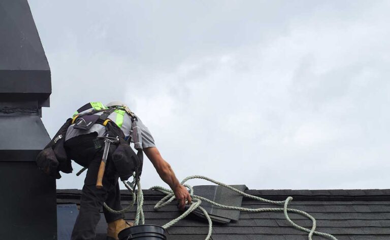 Homeowner inspecting roof shingles from the ground beside a house after winter weather.